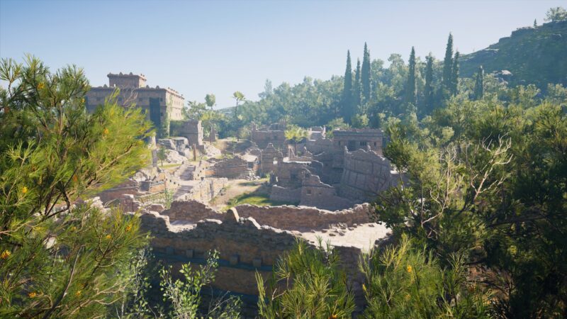 Ruins of ancient stone structures are nestled among lush greenery and tall trees under a clear blue sky, creating a serene and historical atmosphere.