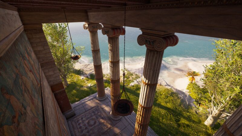 A view from a villa featuring three tall columns, with hanging scales, overlooking a serene beach with white sand and lush greenery by the ocean.