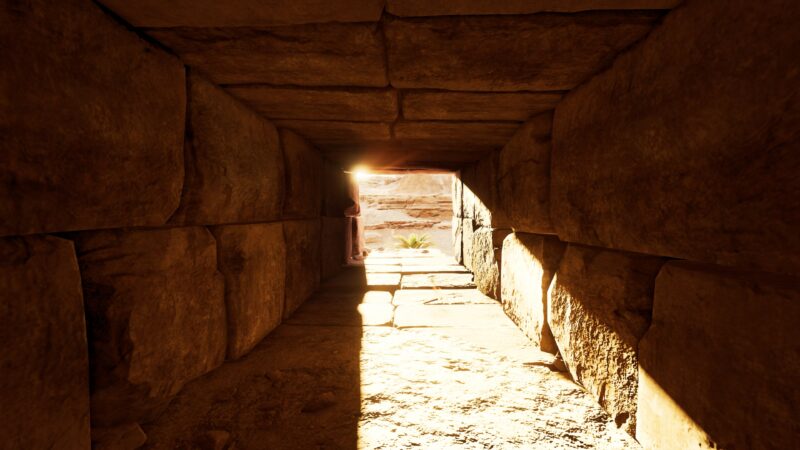 A narrow, stone corridor leads towards bright sunlight illuminating the entrance, revealing textured walls and a sandy floor in an ancient architectural setting.