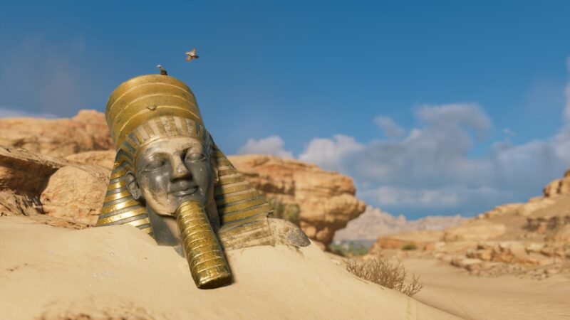 A large, partially buried golden statue of a pharaoh with a headdress sits in a sandy desert landscape under a blue sky, surrounded by rocky formations.