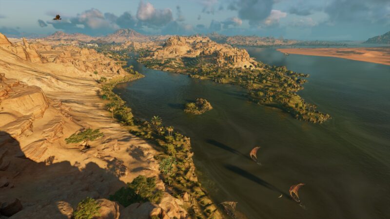 A vast desert landscape features a winding river flanked by lush greenery, with rocky hills in the background and a few sailboats gliding on the water.