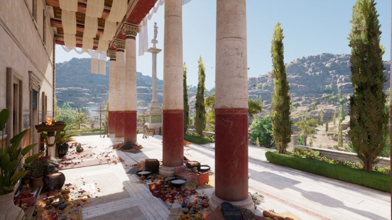 A sunlit ancient courtyard features grand columns, scattered flowers and food offerings, lush greenery, hills in the background, and a statue in the distance.