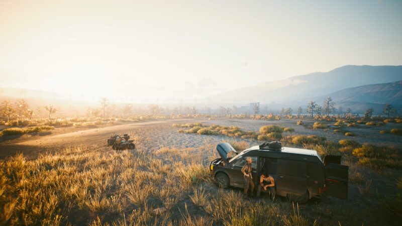 A rugged vehicle sits in a sunlit, sparse landscape with tall grass and distant mountains, while a figure leans against the hood, bathed in warm sunlight.