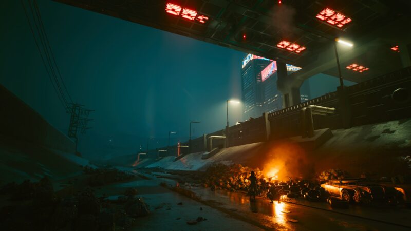 A dimly lit urban scene features a dark alleyway with abandoned cars, piles of debris, and an orange flame illuminating the environment beneath an elevated roadway and towering buildings in the background.