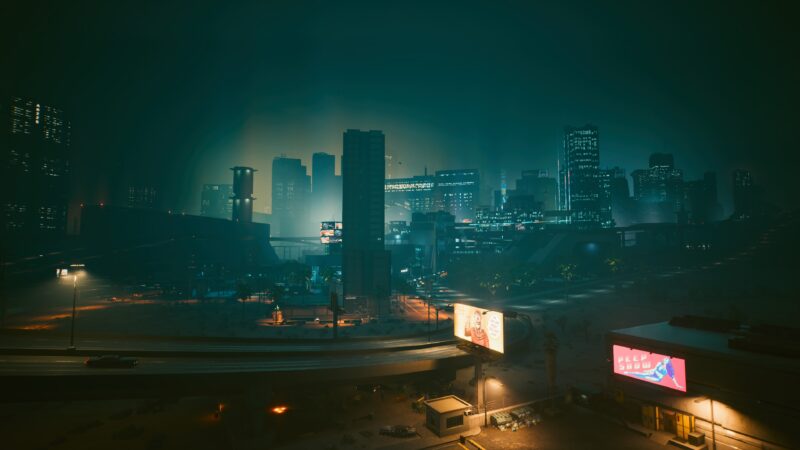 A neon-lit cityscape with towering skyscrapers and hazy atmosphere, featuring a winding road, illuminated billboards, and a blend of industrial architecture set against a darkened sky.