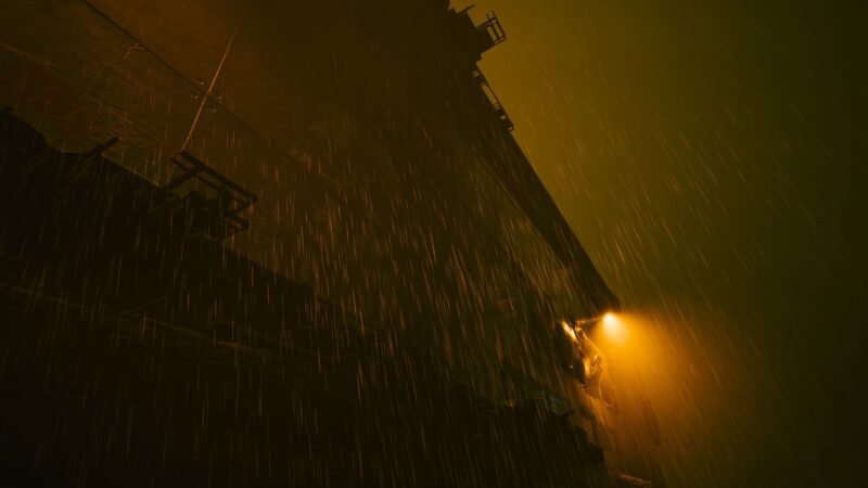 A tall, dark building looms against a misty background, illuminated by a glowing yellow streetlight as heavy rain falls in streaks, creating a moody, atmospheric scene.
