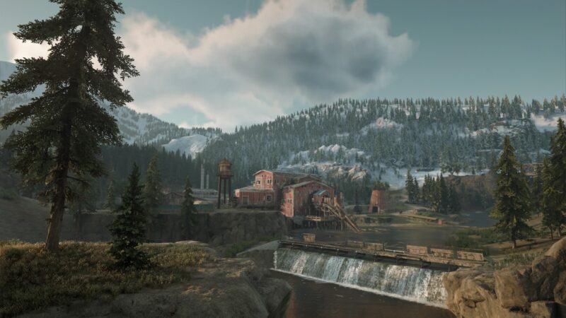 A red wooden structure with multiple buildings overlooks a river flowing over a small dam, surrounded by snow-capped mountains and evergreen trees under a cloudy sky.