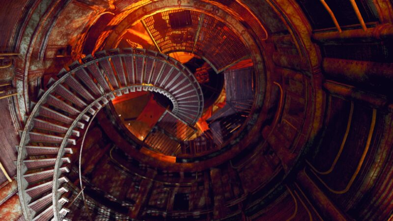 A spiralling metal staircase descends into a rusted, dimly lit circular chamber, illuminated by warm orange light and shadows, with various industrial textures throughout the environment.