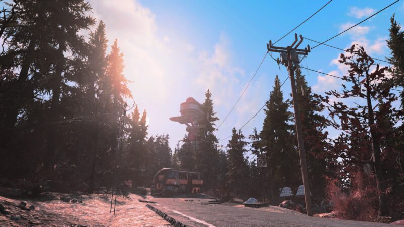 A sunlit road winds through a forest of evergreens, featuring a rusted bus and a distant structure resembling a tower against a vibrant blue sky.