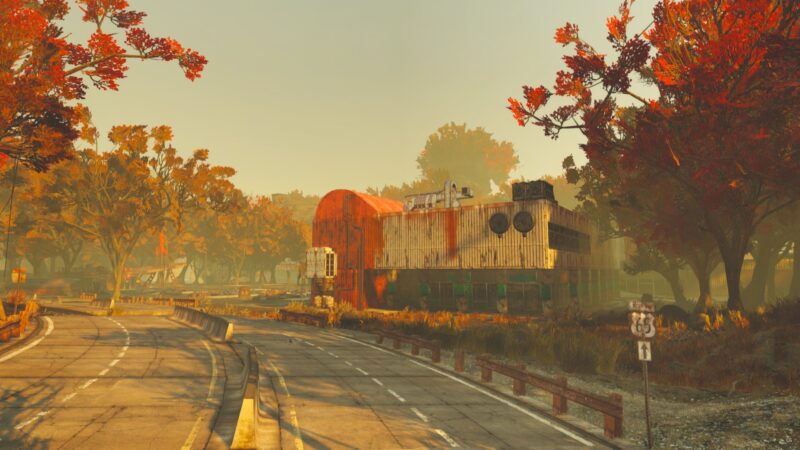 A dilapidated building with a red and white structure stands beside a winding road, surrounded by vibrant autumn trees under a hazy golden sky.