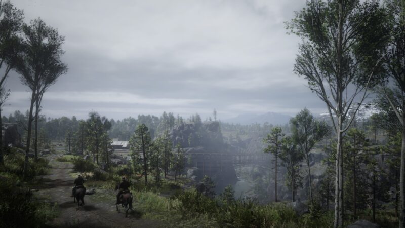 A misty landscape features two riders on horseback along a dirt path, surrounded by lush trees and distant mountains beneath an overcast sky.