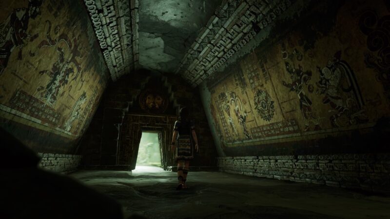 A character walks through a shadowy, ancient temple adorned with detailed murals, revealing a bright exit at the far end, surrounded by stone walls and intricate carvings.