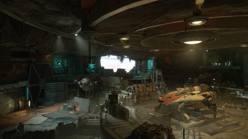 A dimly lit, industrial hangar features a large, orange spaceship on a circular platform, surrounded by various scrap materials and mechanical equipment, with light filtering through a nearby opening.
