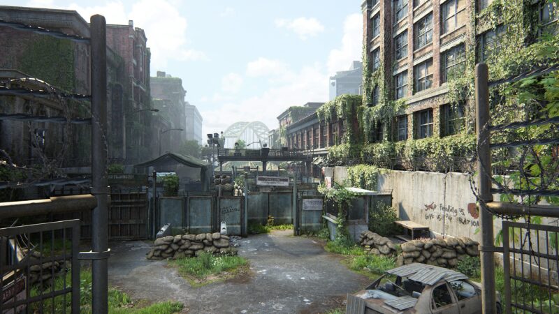 A desolate urban landscape overgrown with vegetation, featuring crumbling buildings, a barricaded entrance, and remnants of structures, under a bright sky scattered with clouds.