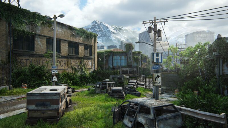 A post-apocalyptic urban landscape features abandoned cars overgrown with grass, crumbling buildings with faded signage, and a distant mountain range under a cloudy sky.