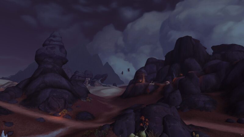 A desolate landscape featuring dark, rocky formations under a stormy sky, with scattered lights along a winding path and hints of distant mountains in the background.