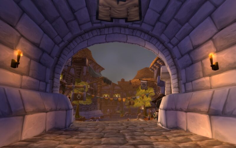 A stone archway frames a cobblestone street bustling with activity, featuring wooden buildings with thatched roofs and banners hanging overhead under a cloudy sky.