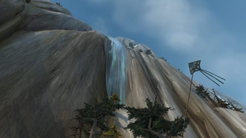 A waterfall cascades down a steep, rocky cliffside surrounded by sparse evergreen trees under a clear blue sky with a distant kite flying.