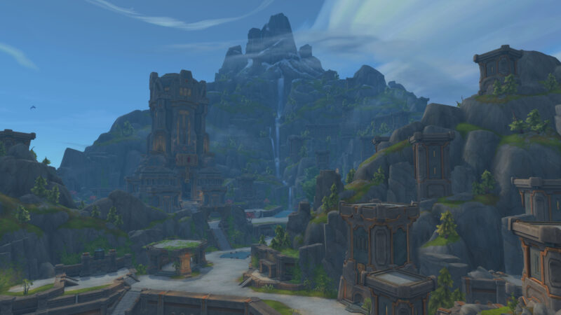A sprawling mountainous landscape features towering stone structures, cascading waterfalls, and lush greenery under a bright blue sky with wispy clouds.